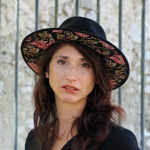 femme brune devant grille avec chapeau noir, dessous en tissus indien motif floral noir rose et vert