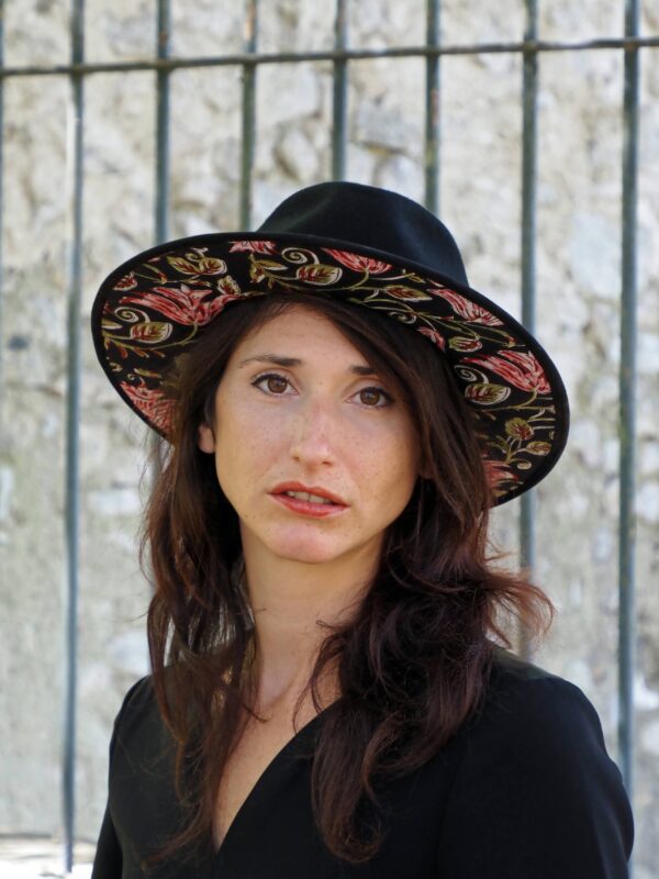 femme brune devant grille avec chapeau noir, dessous en tissus indien motif floral noir rose et vert